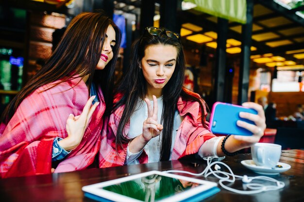 Two young and beautiful girls sitting at the table and doing selfie in the cafe