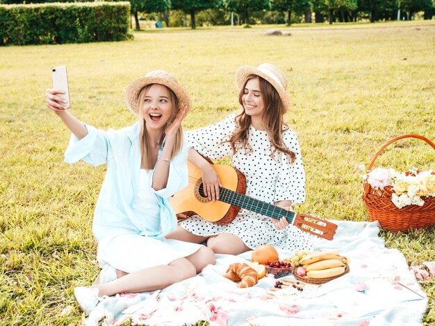 Two young beautiful hipster woman in trendy summer sundress and hats. Carefree women making picnic outside. 