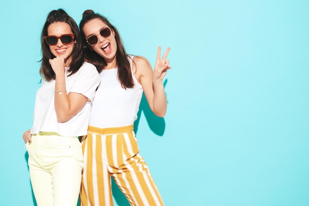 Two young beautiful smiling blond hipster female in trendy summer clothes Sexy carefree women posing near blue wall in studio Positive models having fun Cheerful and happy