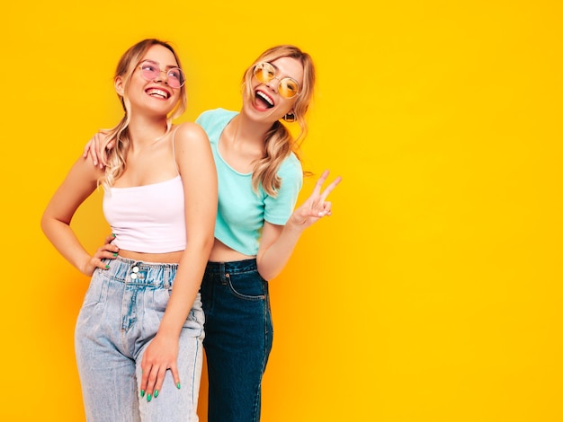 Two young beautiful smiling blond hipster female in trendy summer clothes Sexy carefree women posing near yellow wall in studio Positive models having fun Cheerful and happy In sunglasses