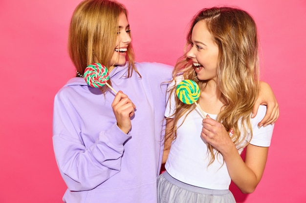 Two young beautiful smiling blond hipster women in trendy summer clothes. Carefree hot women posing near pink wall. Positive funny models with lollipop