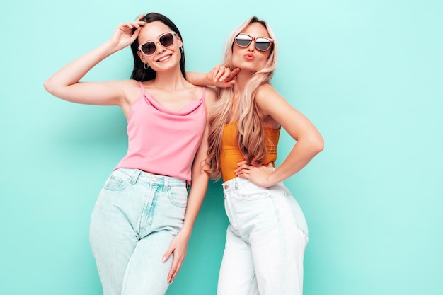 Two young beautiful smiling brunette hipster female in trendy summer clothes Sexy carefree women posing near blue wall Positive models having fun Cheerful and happy