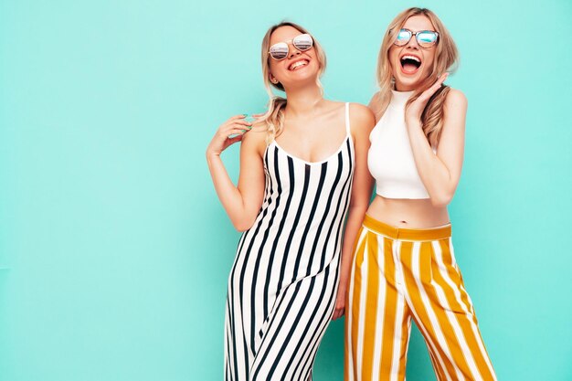 Two young beautiful smiling brunette hipster female in trendy summer clothes Sexy carefree women posing near blue wall in studio Positive models having fun Cheerful and happy