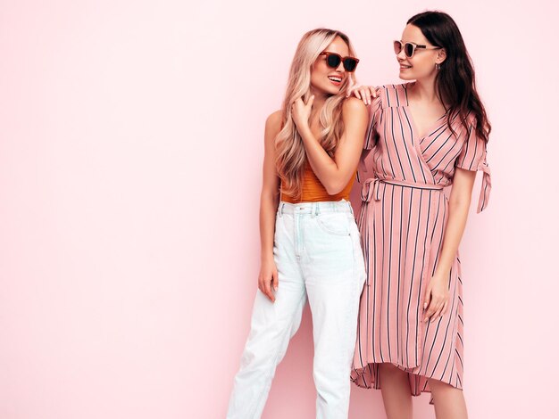 Two young beautiful smiling brunette hipster female in trendy summer clothes Sexy carefree women posing near pink wall Positive models having fun Cheerful and happy In sunglasses