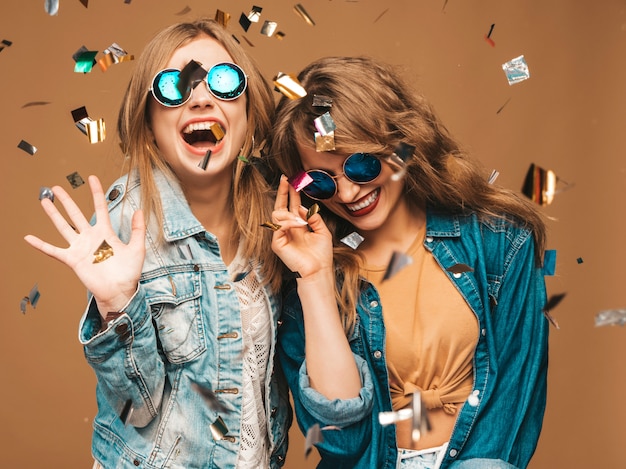 Two young beautiful smiling girls in trendy summer clothes and sunglasses. Sexy carefree women posing. Positive screaming models under confetti