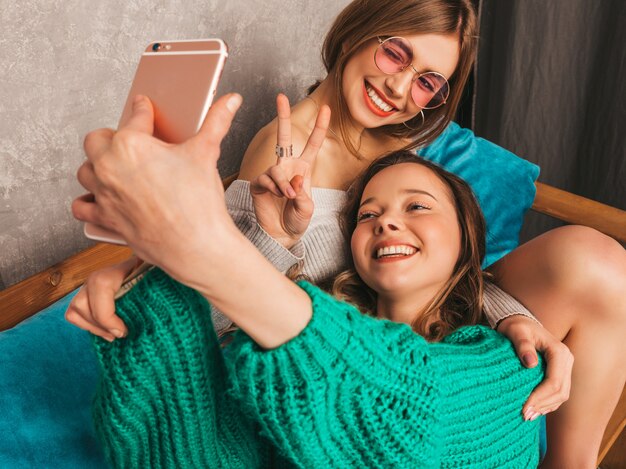 Two young beautiful smiling gorgeous girls in trendy summer clothes.  Sexy carefree women posing in interior and taking selfie. Positive models having fun with smartphone