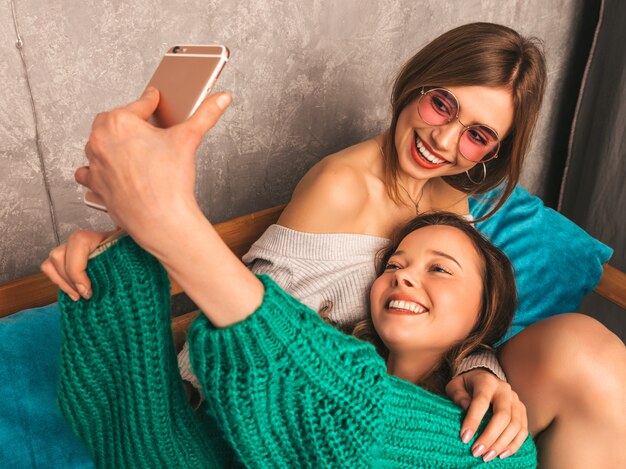 Two young beautiful smiling gorgeous girls in trendy summer clothes.  Sexy carefree women posing in interior and taking selfie. Positive models having fun with smartphone