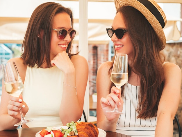 Two young beautiful smiling hipster female in trendy summer clothesCarefree women posing at veranda cafe in the streetPositive models drinking white vine in hatEnjoying their vacation