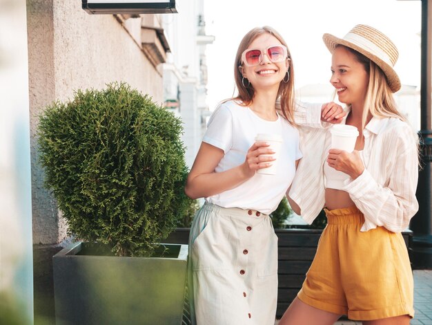 Two young beautiful smiling hipster female in trendy summer clothesSexy carefree women posing in the street Positive pure models having fun at sunset They drinking coffee or tea in plastic cup