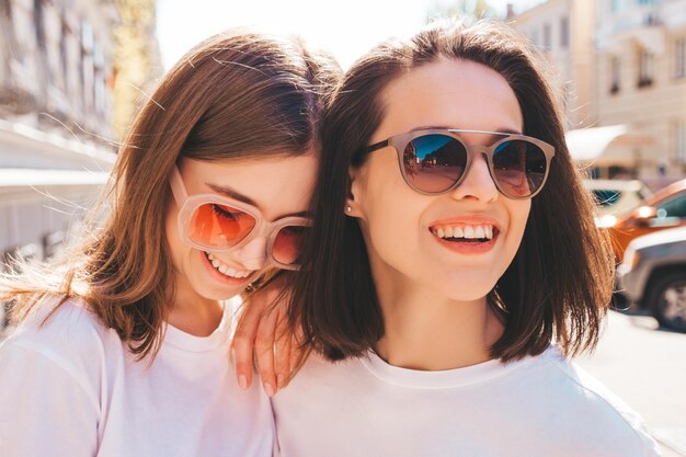 Two young beautiful smiling hipster female in trendy summer white t-shirt clothes and jeans.Sexy carefree women posing on the street background. Positive models having fun, hugging and going crazy