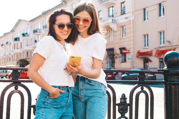 Two young beautiful smiling hipster female in trendy summer white t-shirt clothes and jeans.Sexy carefree women posing on the street background.Positive models using smartphone apps, looking at screen