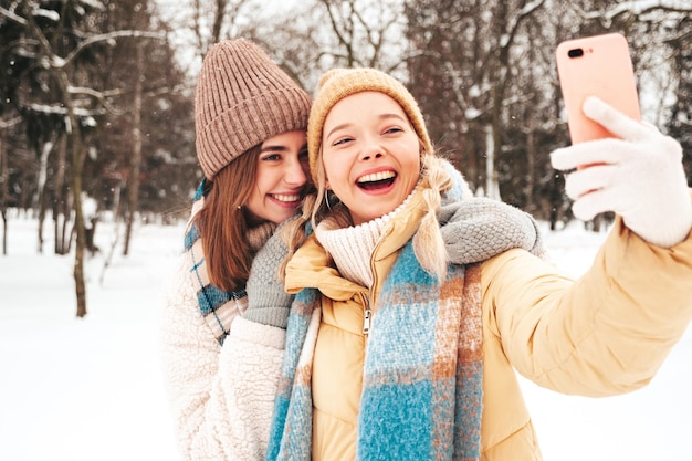 Free Photo two young beautiful smiling hipster female in trendy warm clothes and scarfs.carefree women posing in the street in park. positive pure models having fun in snow. enjoying winter moments.taking selfie