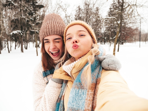 Two young beautiful smiling hipster female in trendy warm clothes and scarfs.Carefree women posing in the street in park. Positive pure models having fun in snow. Enjoying winter moments.Taking selfie