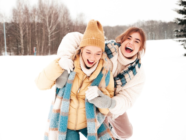 Free Photo two young beautiful smiling hipster female in trendy warm clothes and scarfs.carefree women posing in the street in park. positive pure models having fun in snow. enjoying winter moments. xmas concept