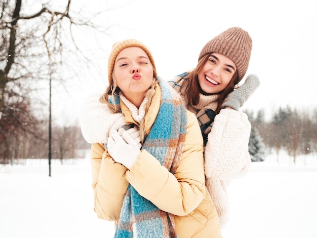 Free Photo two young beautiful smiling hipster female in trendy warm clothes and scarfs.carefree women posing in the street in park. positive pure models having fun in snow. enjoying winter moments. xmas concept