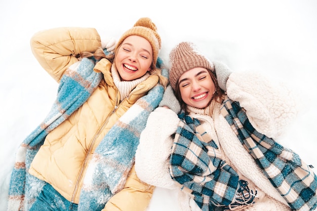 Free Photo two young beautiful smiling hipster female in trendy warm clothes and scarfs.carefree women posing in the street in park. positive pure models lying in snow. enjoying winter moments. xmas concept