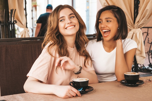 Free photo two young beautiful smiling hipster girls in trendy summer clothes.carefree women chatting in veranda terrace cafe and drinking coffee.positive models having fun and communicating