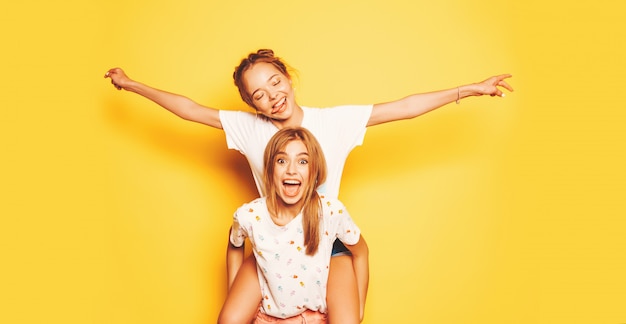 Two young beautiful smiling hipster girls in trendy summer clothes. Sexy carefree women posing near yellow wall.Model sitting on her friend's back and raising hands