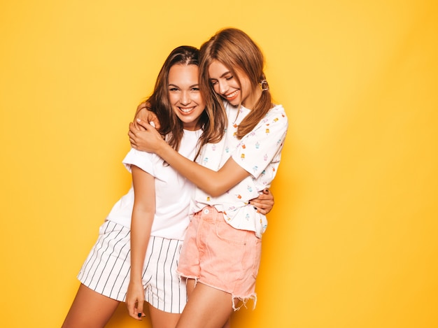 Free photo two young beautiful smiling hipster girls in trendy summer clothes. sexy carefree women posing near yellow wall. positive models going crazy and having fun.hugging