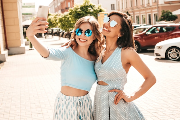 Two young beautiful smiling hipster girls in trendy summer clothes.Sexy carefree women posing on the street background in sunglasses. They taking selfie self portrait photos on smartphone at sunset