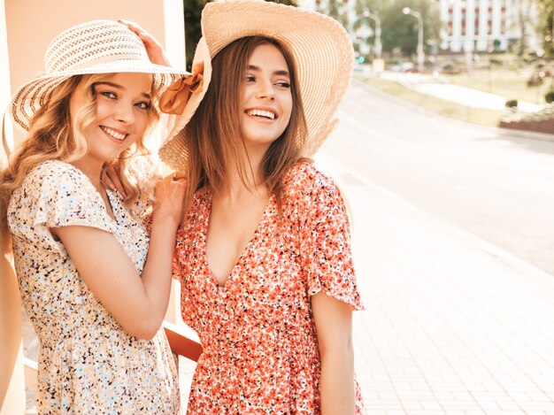 Two young beautiful smiling hipster girls in trendy summer sundress.Sexy carefree women posing on the street background in hats. Positive models having fun and hugging