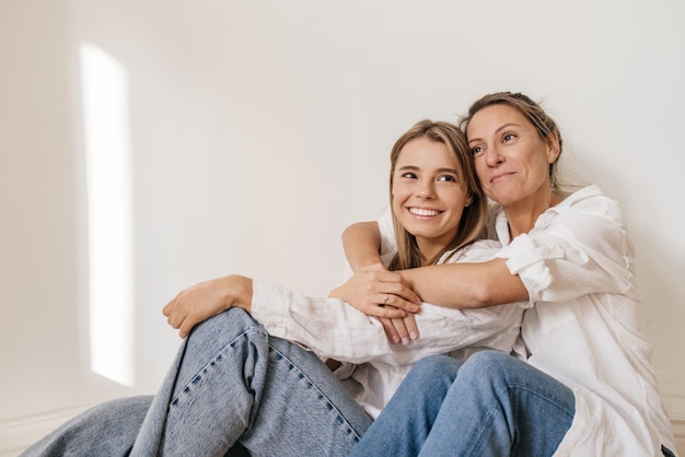 Two young caucasian women wear casual clothes, sit on floor against white wall and enjoy spending time together. Leisure, lifestyle concept