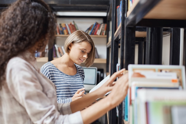 Two young charming students girls standing near bookshelf, looking for information for group project in library, talking about points of team work.