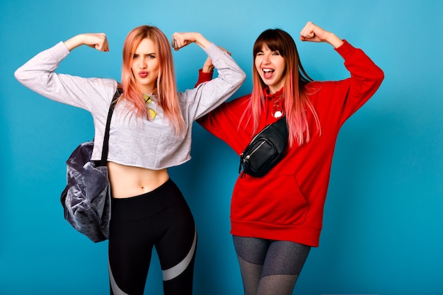 Two young funny pretty hipster women wearing sportive bright casual outfits, showing biceps and making grimaces, going crazy together,  blue wall