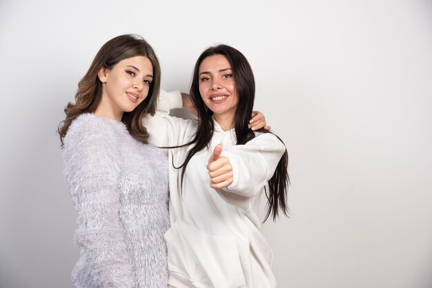 Two young girl friends smiling and showing a thumb up on white wall .