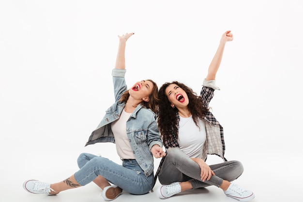 Two young girls sitting on the floor together while screaming and looking up over white wall