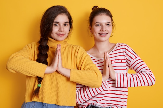 Free photo two young girls wearing casual shirts posing with palms together and smiling