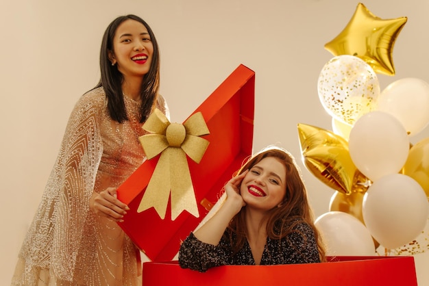 Two young interracial girls redhead and brunette are smiling at camera against backdrop of balloons and gift red big box