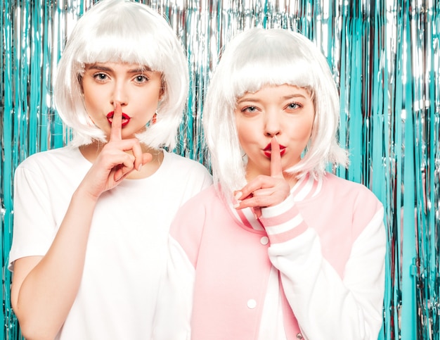 Free Photo two young sexy hipster girls in white wigs and red lips.beautiful trendy women in summer clothes.models posing on blue silver shiny tinsel background in studio.showes finger hush silence sign, gesture