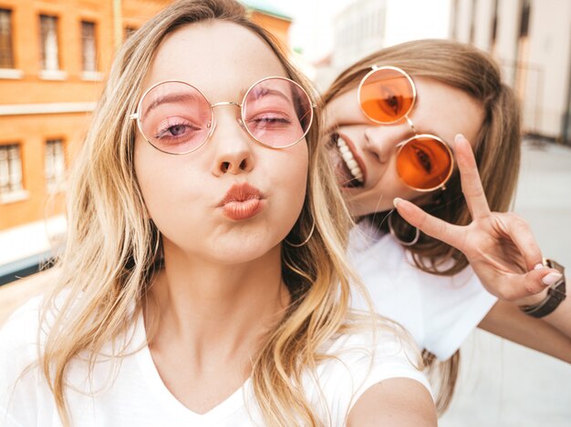 Two young smiling hipster blond women in summer clothes. Girls taking selfie self portrait photos on smartphone. . 