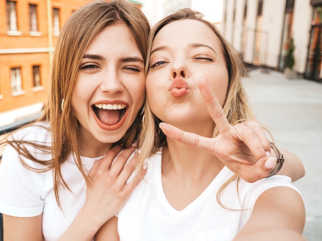 Two young smiling hipster blond women in summer clothes. Girls taking selfie self portrait photos on smartphone. . 