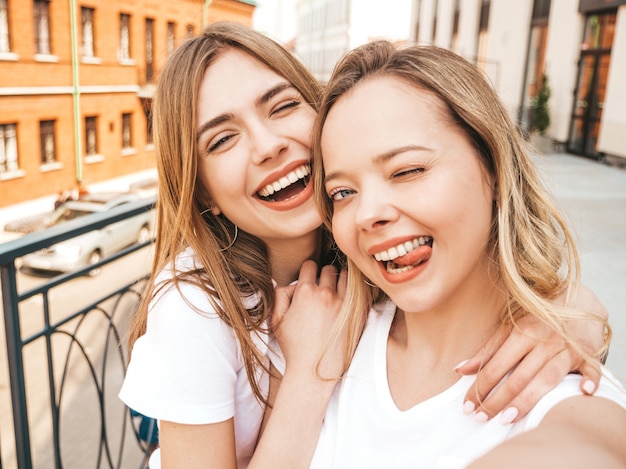 Free photo two young smiling hipster blond women in summer white t-shirt clothes. girls taking selfie self portrait photos on smartphone.  .female showing tongue