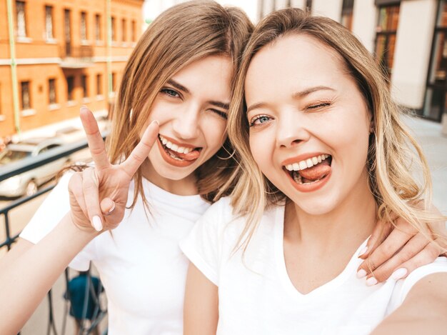 Two young smiling hipster blond women in summer white t-shirt clothes. Girls taking selfie self portrait photos on smartphone.  .Female shows peace sign and tongue