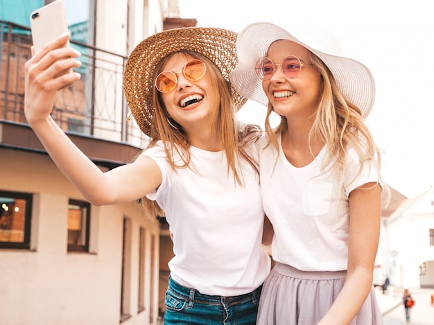 Two young smiling hipster blond women in summer white t-shirt clothes. Girls taking selfie self portrait photos on smartphone. 