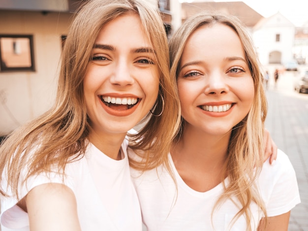 Two young smiling hipster blond women in summer white t-shirt clothes. Girls taking selfie self portrait photos on smartphone. 