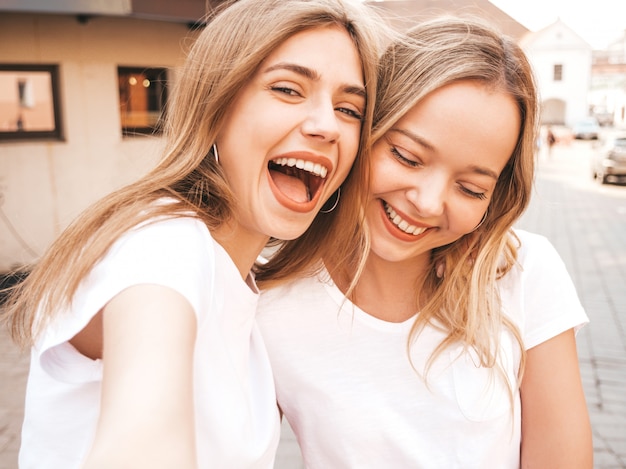 Two young smiling hipster blond women in summer white t-shirt clothes. Girls taking selfie self portrait photos on smartphone. 