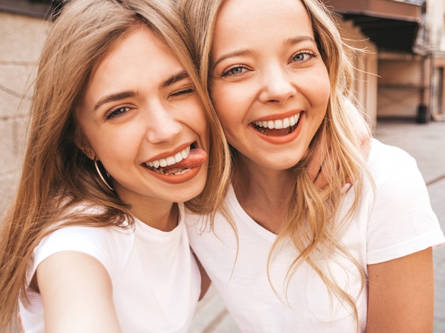 Two young smiling hipster blond women in summer white t-shirt clothes. Girls taking selfie self portrait photos on smartphone. 