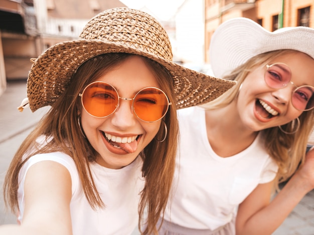 Free Photo two young smiling hipster blond women in summer white t-shirt. girls taking selfie self portrait photos on smartphone.models posing on street background.female shows tongue and positive emotions