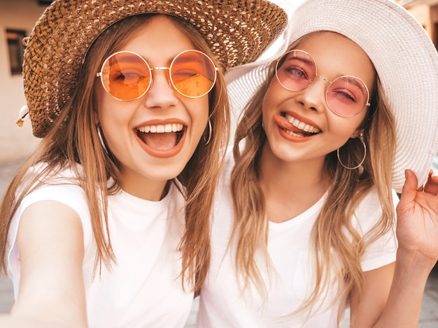 Free photo two young smiling hipster blond women in summer white t-shirt. girls taking selfie self portrait photos on smartphone.models posing on street background.female shows tongue and positive emotions