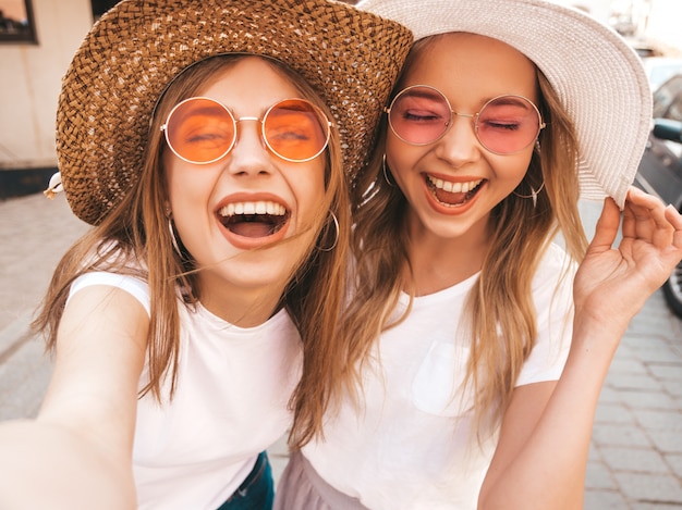 Free photo two young smiling hipster blond women in summer white t-shirt. girls taking selfie self portrait photos on smartphone.models posing on street background.female shows tongue and positive emotions