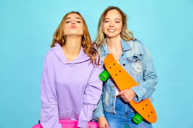 Free photo two young stylish smiling blond women with penny skateboards. women in summer hipster sport clothes posing near blue wall . positive models