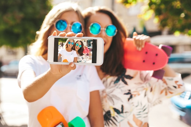 Two young stylish smiling hippie brunette in summer sunny day in hipster clothes with penny skateboard posing