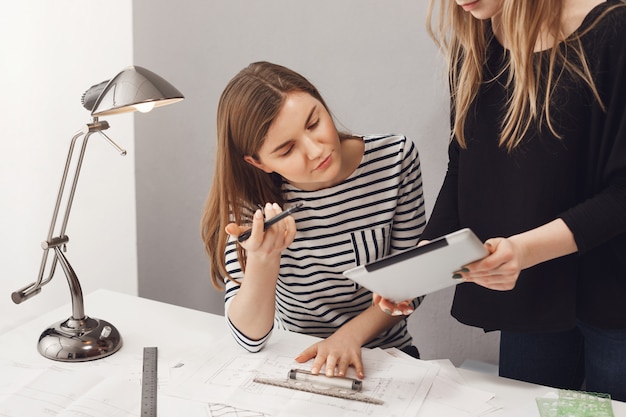 Two young successful freelance female architects looking through working plan, talking about deadlines, making blueprints for customer. Freelance, business and teamwork concept.