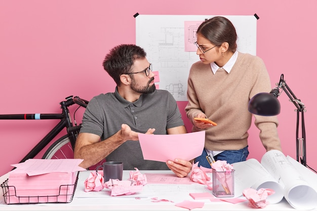 Free photo two young woman and man coworkers look angrily at each other blame for mistake pose at modern office desk discuss sketch for building project. professional engineers collaborate on blueprints