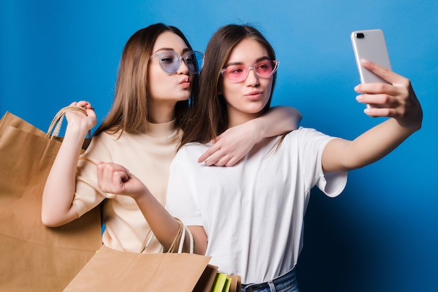 Free Photo two young women take selfie on the phone with colorful paper bags isolated on blue wall. concept for shop sales.