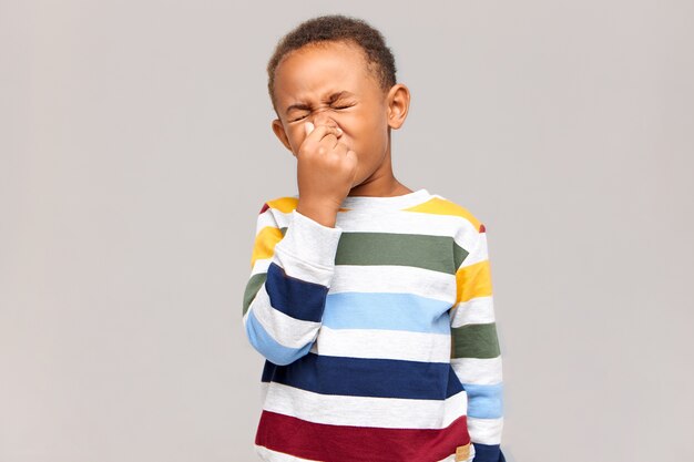 Ugh, disgusting! Portrait of emotional disgusted African American boy closing eyes and pinching nose because of bad smell or stink.  dark skinned male child having allergy, sneezing